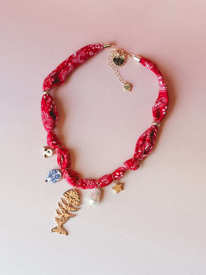 A red bandana-print fabric choker necklace laid flat, strung with an eclectic mix of charms — a gold fish skeleton, baroque pearl, star, ceramic coin bead, and small heart — all dangling from a knotted, twisted scarf-style band with a silver extender