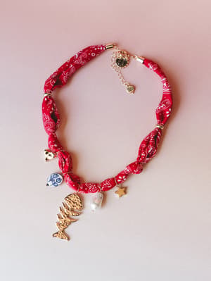 A red bandana-print fabric choker necklace laid flat, strung with an eclectic mix of charms — a gold fish skeleton, baroque pearl, star, ceramic coin bead, and small heart — all dangling from a knotted, twisted scarf-style band with a silver extender