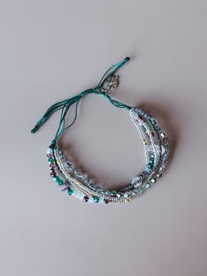 A multi-strand beaded bracelet stack laid flat against a soft gray background, mixing crystal, seed bead, and faceted glass strands in teal, silver, gray, and mixed jewel tones — finished with an adjustable teal cord closure and a small silver sun ch