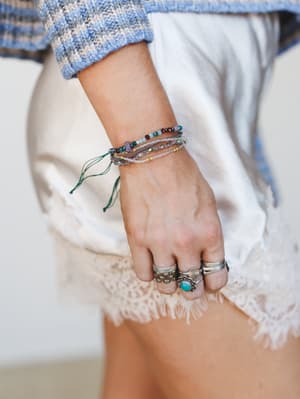 A close-up of stacked beaded bracelets and silver rings styled on a model's hand, layered with mixed-stone bracelets in earthy tones against a soft white and blue knit background — the kind of jewelry stack that looks like it was collected, not coord