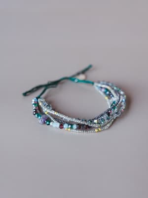 A multi-strand beaded bracelet stack laid on a soft gray surface, mixing seed beads, faceted crystals, and a raw stone accent in a cool, jewel-toned palette tied with a teal cord closure.