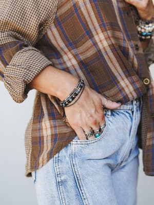 A close-up lifestyle shot focused on a stacked beaded bracelet worn at the wrist, styled with a plaid flannel shirt, gingham cuffs, light wash jeans, and a collection of silver and turquoise rings — the kind of effortless accessory layering that look