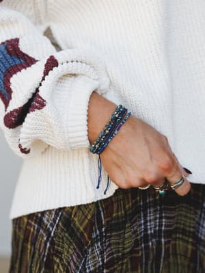 A close-up of a stacked beaded bracelet set in deep blue and teal layered on a wrist, styled against a chunky cream knit sweater sleeve and a plaid skirt — the kind of detail shot that makes you reach for your own wrist to see what's there.