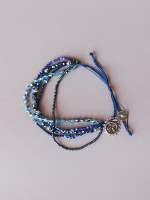 A multi-strand beaded bracelet stack in layered blues — seed beads, faceted crystals, and dark hematite-style beads gather at an adjustable braided cord closure hung with a silver peace sign charm and a small starburst coin charm.