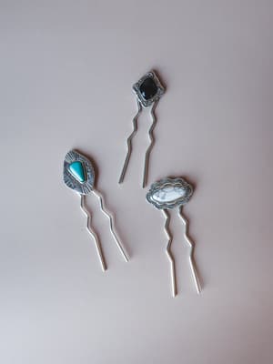 Three silver hair pins laid flat on a muted mauve surface, each topped with a different stone — turquoise, black onyx, and white howlite — with stamped southwestern-style metalwork framing each one.