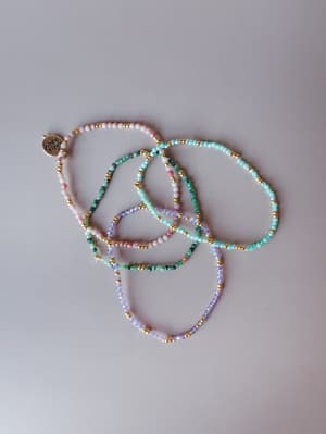 A loose, casually arranged set of delicate seed bead bracelets in soft pastel and jewel tones with gold accent beads, photographed flat on a light grey surface — the kind of jewelry stack that looks like it was made for a slow summer day.