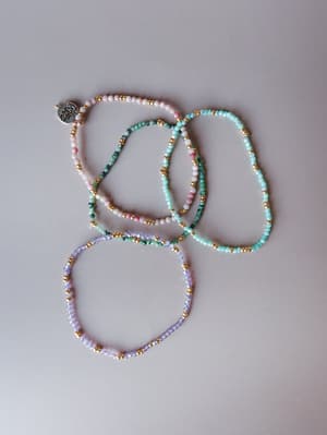 A loose, casually arranged set of delicate seed bead bracelets in soft pastel tones with gold accent beads, laid flat on a light grey surface — the kind of jewelry that ends up on your wrist every single day without you thinking twice.