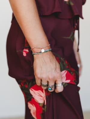A close-up of a stacked beaded bracelet set on a model's wrist, layered with silver rings including a turquoise stone — all set against a rich burgundy floral dress that makes the mixed-color beads pop.