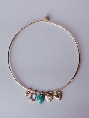A delicate gold wire choker collar necklace laid flat against a soft gray background, strung with an eclectic mix of charms — a faceted crystal, a green stone drop, a sunburst heart, a puffed heart, and a tiny textured charm — the kind of piece that