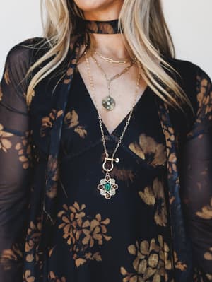 A close-up styled shot showing layered gold necklaces — including a statement gem pendant and coin charm — against a sheer black and golden floral dress with a deep v-neckline and black choker, feeling rich and a little mysterious.