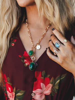 A close-up of layered gold chain necklaces with deep green oval stone pendants and a coin charm, styled over a burgundy floral top with stacked boho rings — the kind of jewelry that looks like it was collected, not purchased.