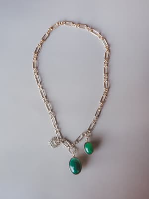 A silver paperclip chain necklace laid flat, styled with three charms — two oval green stone pendants and a small starburst coin charm — shot clean against a soft grey surface.