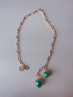 A gold paperclip chain necklace laid flat on a neutral grey surface, featuring two oval green stone pendants of different sizes and a small starburst charm — the kind of piece that feels like it was found, not bought.