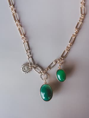 A gold paperclip-chain charm necklace laid flat, featuring two oval green cabochon pendants, a crystal-pavé connector link, and a silver sunburst coin charm — the kind of piece that looks like it was collected over time, not bought all at once.