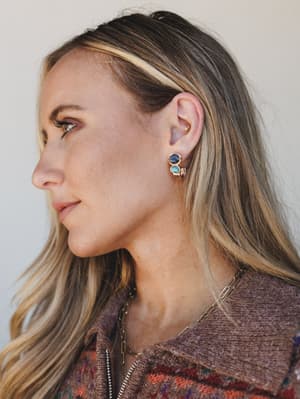 A close profile shot highlighting a stacked stone earring in gold with deep blue and turquoise cabochon details, styled against a soft neutral background with a cozy mauve knit and layered chain necklace peeking into frame.