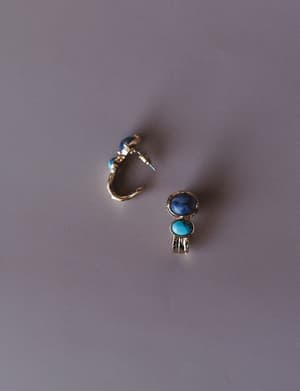 A pair of gold hoop earrings set with dual oval stone cabochons — one deep sodalite blue, one turquoise — photographed flat against a muted lavender-gray surface, showing both the front face and the open hoop back.