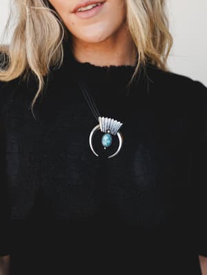 A close-up shot of a statement southwestern-style pendant necklace — silver crescent horn with a turquoise stone center on a black cord, worn over a black floral-textured top.
