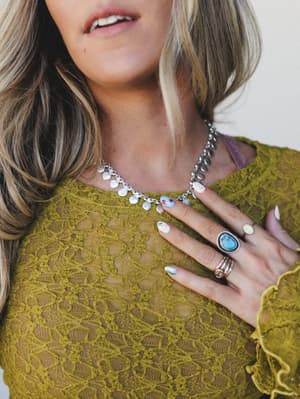 A close-up shot of a model in a olive lace top, hand resting on her chest to show off a boho silver coin charm necklace alongside a turquoise stone ring and painted nails — the kind of jewelry stack that feels personal, not put-together.