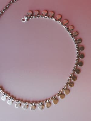 A silver coin charm necklace laid flat on a dusty rose background — the kind of piece that quietly pulls an outfit together without trying too hard.