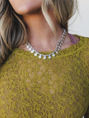 A close-up detail shot highlighting a silver coin-charm necklace layered over a sheer olive lace top, the combo feeling effortlessly collected and just a little earthy-romantic.