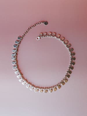 A silver coin charm necklace laid flat against a dusty rose background — the kind of piece that sounds like movement and somehow makes a plain white tee feel intentional.