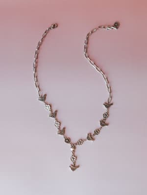 A silver chain necklace laid flat against a soft pink gradient background, featuring alternating bird and daisy charms with an Y-drop pendant — the kind of piece that feels like it has a little story in it.
