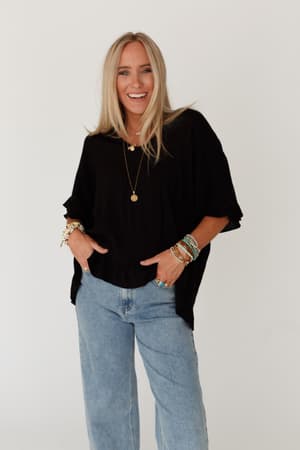 A smiling woman with long blonde hair wears The Nest Let Me Be Ruffled Top in black, light blue jeans, layered necklaces, and colorful bracelets, standing against a plain white background.