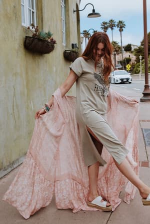A woman with long red hair, smiling on a sidewalk, wears a beige T-shirt dress and white slides. She holds out the Paisley Tapestry Free Flow Duster Kimono in Blush for a breezy look, accented by layered bracelets.