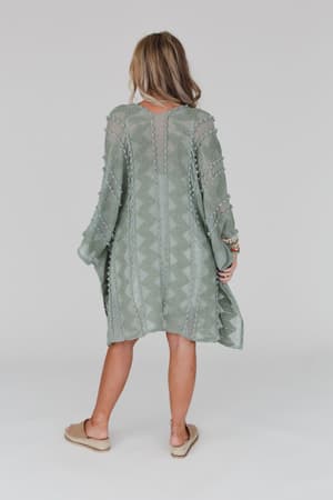 A woman with shoulder-length hair, sandals, and bracelets wears the Bali Embroidered Kimono in sage—a loose, textured piece with geometric patterns—standing against a plain, light-colored background.