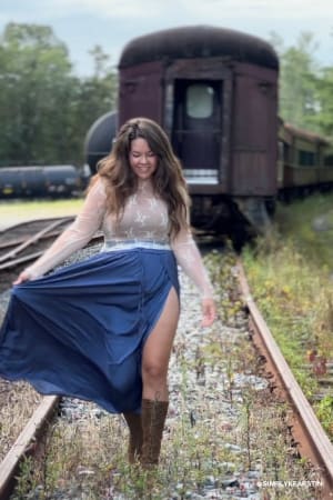Woman in a blue skirt and lace top standing on train tracks with a vintage train in the background