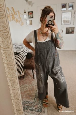 A woman takes a mirror selfie in a cozy room, wearing the High Hopes Slouchy Denim Overalls - Black Bandana over a white tee, paired with sandals and jewelry. The space features wall art, a rug, and a zebra-striped bench.