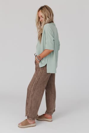 A woman with long, wavy blonde hair wears a light sage green top, So Comfy Wide Leg Pant Full Length in Mocha, beige espadrille sandals, and colorful bracelets, posing against a plain white background.