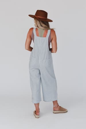 A woman stands facing away, wearing a brown hat, a light top, and the High Hopes Denim Overalls in Denim Stripe. Her hair is down and she’s on a plain white background in sandals for a chic look.