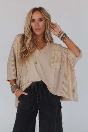 A woman with long blonde hair wears the City Drifter Oversized Contrast Top in taupe, styled with black drawstring pants, layered necklaces, and colorful beaded bracelets against a plain light background.