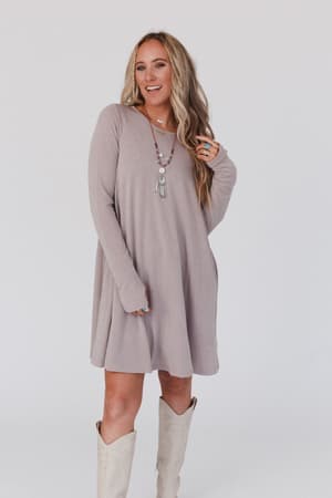 A woman with long blonde hair wears The Nest Jake Long Sleeve Crewneck Dress in Mocha Latte and knee-high cream boots, accessorized with layered necklaces and rings, smiling against a plain white background.