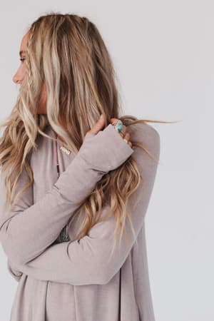 A woman with long, wavy blonde hair wears The Nest Jake Long Sleeve Crewneck Dress in Mocha Latte. She looks left with arms crossed, one hand touching her hair, displaying a turquoise ring and layered necklaces against a plain white background.
