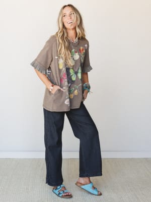 A laughing model styled in an oversized ash brown butterfly graphic tee with ruffle sleeves, paired with dark wide-leg jeans and sky blue Birkenstock sandals — the kind of effortless outfit that looks like you meant to do that.