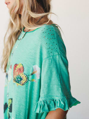 A close-up detail shot of a teal green top or dress with a butterfly graphic, eyelet cutout shoulder panel, and ruffled sleeve hem — the kind of piece that feels like it was made for a slow morning somewhere warm.