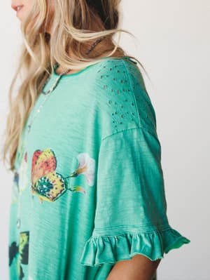 A close-up detail shot of a teal green top or dress with a butterfly graphic, eyelet cutout shoulder panel, and ruffled sleeve hem — the kind of piece that feels like it was made for a slow morning somewhere warm.
