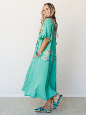 A flowy mint green maxi dress with a botanical butterfly print and ruffle sleeve detail, styled with stacked bracelets and crochet sandals against a clean studio wall — effortless and a little whimsical.