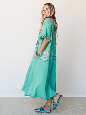 A flowy mint green maxi dress with a botanical butterfly print and ruffle sleeve detail, styled with stacked bracelets and crochet sandals against a clean studio wall — effortless and a little whimsical.