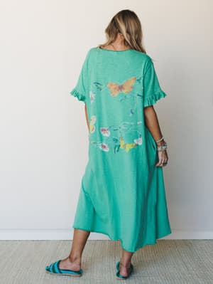 A breezy kelly green maxi dress photographed from the back, with a whimsical butterfly and floral graphic wrapping across the silhouette — the kind of dress that makes you want to wander somewhere with flowers in it.