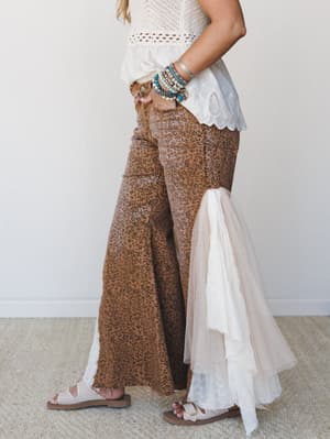 A side-profile shot of wide-leg leopard print pants in warm camel tones, styled with a crochet and eyelet top, layered boho bracelets, and sandals — the ruffle lace hem detail at the ankle gives the whole look an unexpected, free-spirited finish.