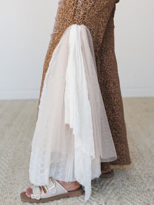 A close-up side view of wide-leg leopard print pants in warm camel tones, styled over a sheer blush tulle and eyelet underlayer, finished with cream buckle sandals — the kind of unexpected mix that feels effortlessly personal.