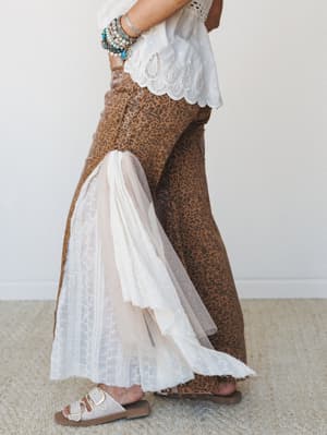 A side-profile shot showing wide-leg leopard print pants in warm camel tones layered with an eyelet lace hem insert and a flowing lace panel — styled with a white eyelet top, stacked beaded bracelets, and neutral buckle sandals for a look that feels