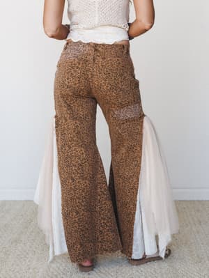 Back view of wide-leg leopard print pants in warm camel with dramatic tulle and lace side panels that flare out at the hem, paired with a cream crochet crop top — the kind of outfit that stops a room without trying.