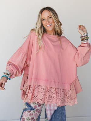 A smiling model in a roomy dusty rose sweatshirt-style top with layered eyelet and ruffle hem details, styled with patchwork jeans and stacked beaded bracelets — the kind of outfit that feels effortless and still totally put-together.