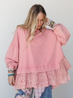 A relaxed, oversized dusty rose top with layered eyelet ruffle hem worn with patchwork wide-leg pants and a stack of colorful bracelets — the kind of outfit that feels effortless but very much on purpose.