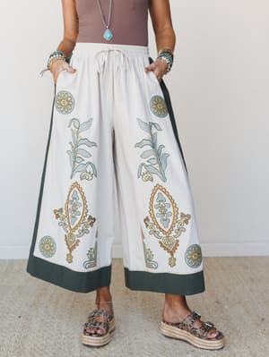 Wide-leg pants in cream with bold botanical and medallion block print detailing, forest green contrast border at hem and side panels, styled with a mauve bodysuit, turquoise jewelry, and leopard espadrille sandals — effortlessly boho with a well-trav