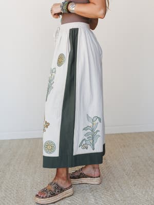 A side-angle shot of wide-leg pants in cream and forest green with botanical block-print embroidery, styled with stacked turquoise bracelets and leopard platform sandals — earthy and expressive in the best way.